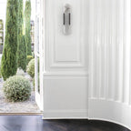 The Clérachic sconce unlit on a white panelled wall, framed by open garden doors and clipped cypress