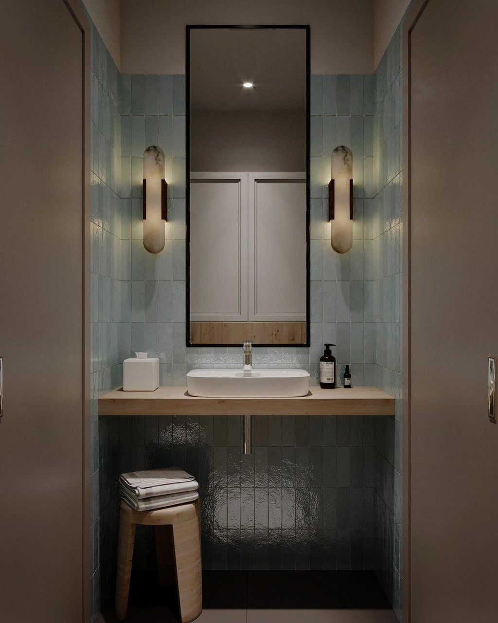 Two Clérachic sconces flanking a slim mirror in a blue-green tiled powder room above the basin and oak stool