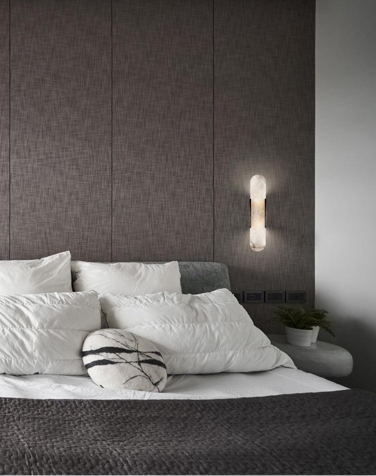 Clérachic sconce lit beside a bed dressed in white linen, against a taupe fabric-panelled headboard wall