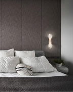 Clérachic sconce lit beside a bed dressed in white linen, against a taupe fabric-panelled headboard wall