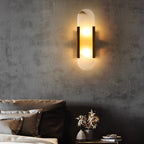 The Clérachic sconce lit on a raw concrete bedroom wall above taupe pillows and dried stems