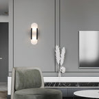 The Clérachic sconce with black bars lit on a grey panelled wall above a green chair and marble ledge