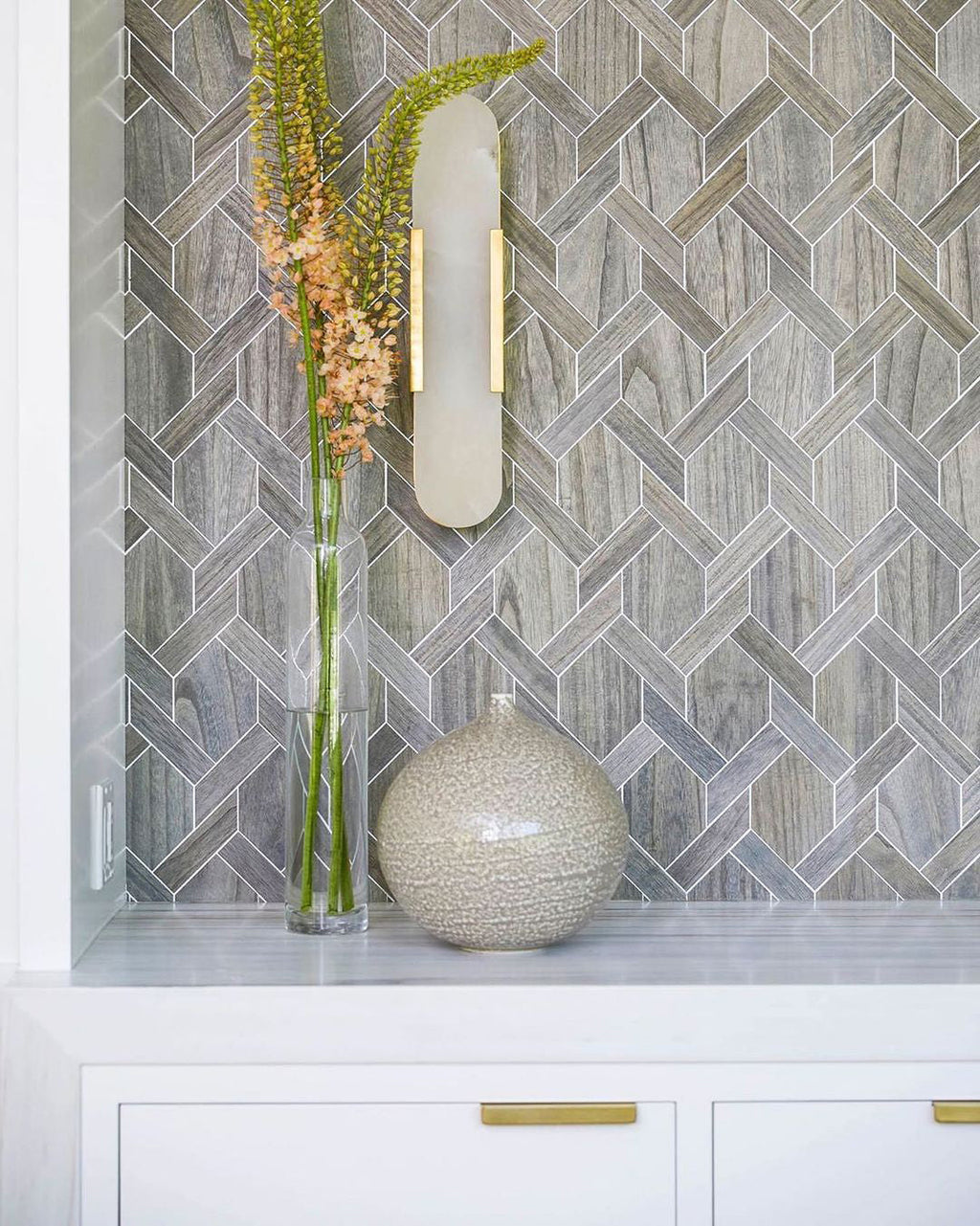 Clérachic sconce lit on a grey mosaic tiled niche, a glass vase of blooms on the ledge below