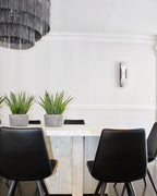 The Clérachic sconce unlit on a white dining-room wall beside potted greenery and black chairs