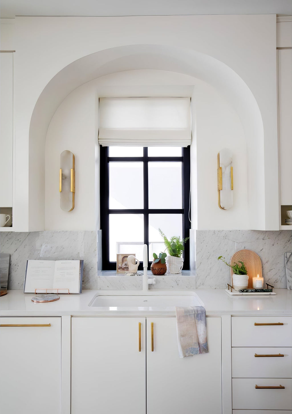 Clérachic sconces set either side of an arched kitchen window above white cabinetry and brass handles