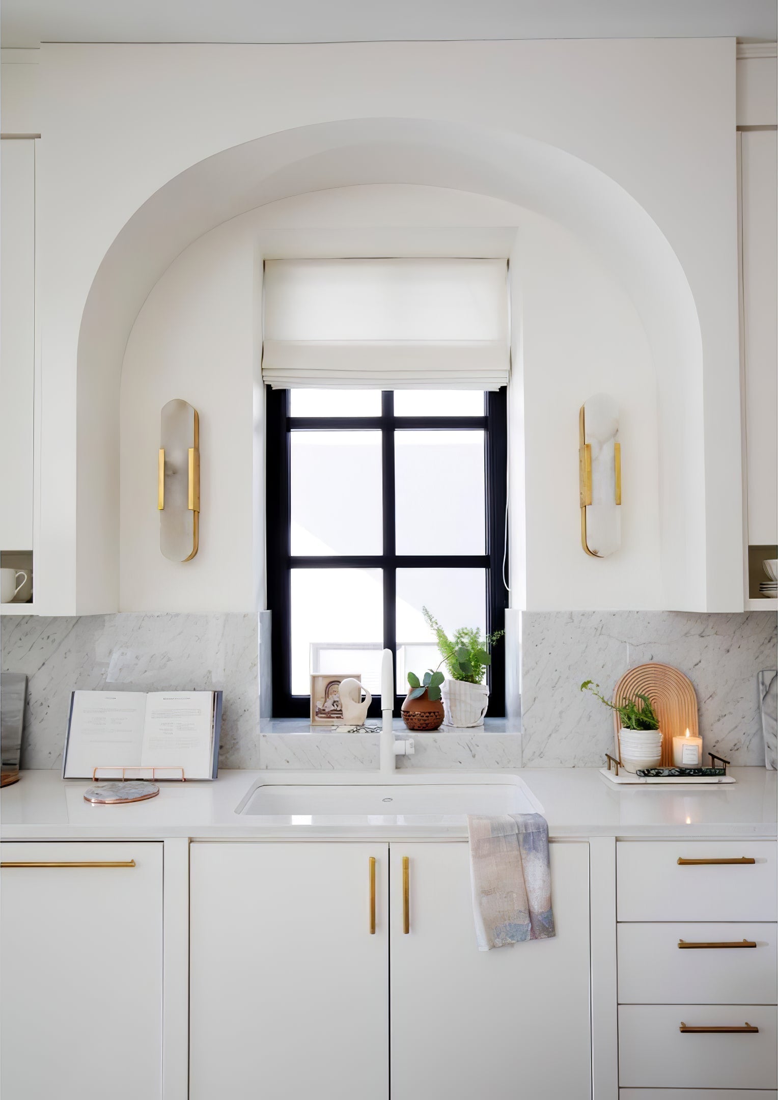 Clérachic sconces set either side of an arched kitchen window above white cabinetry and brass handles
