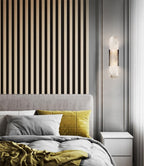 Clérachic sconce lit on a grey wall beside a vertical oak slat panel and a bed in grey and mustard
