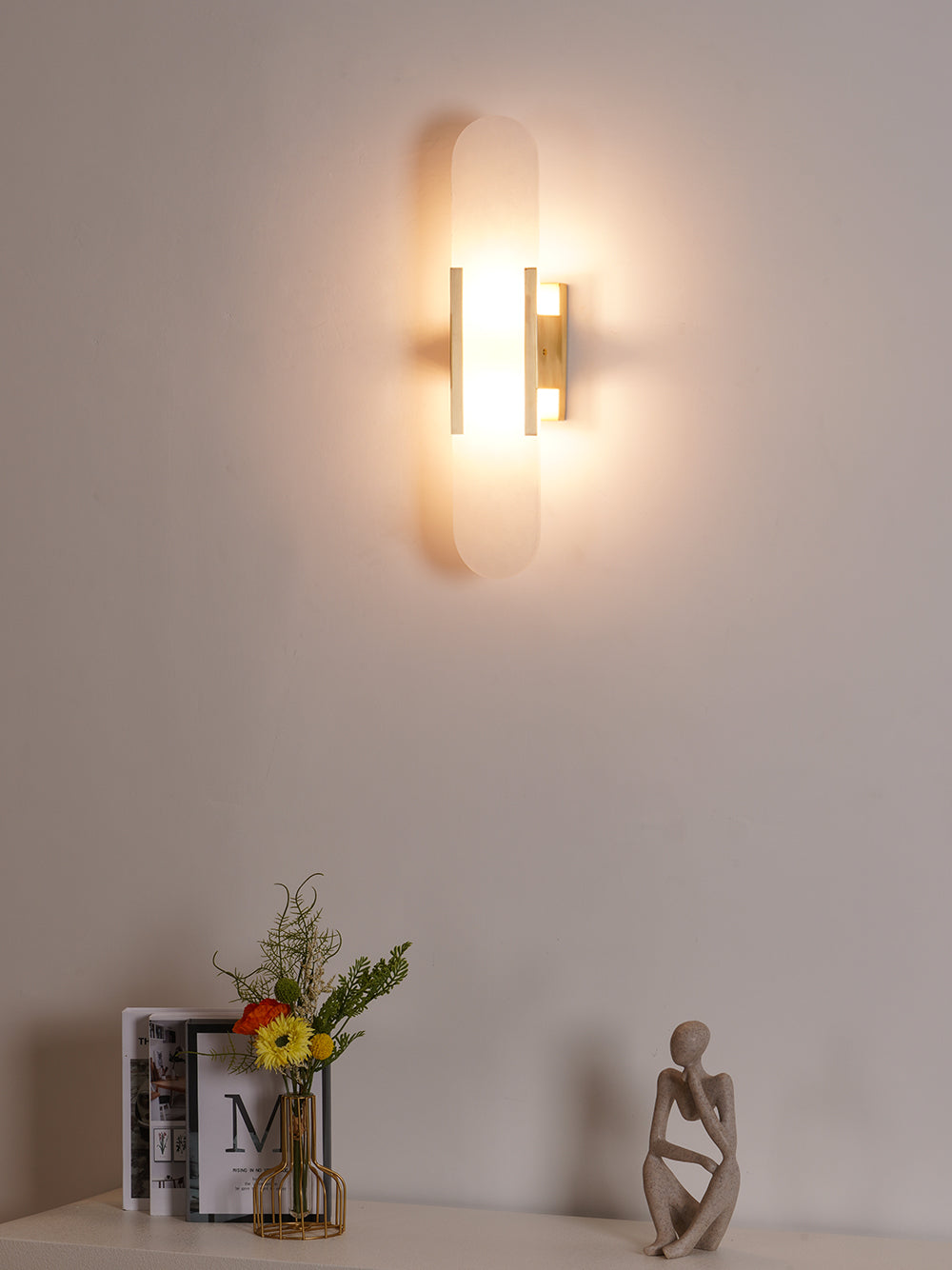 The Clérachic sconce lit on a pale wall above a shelf of flowers, books and a carved figure