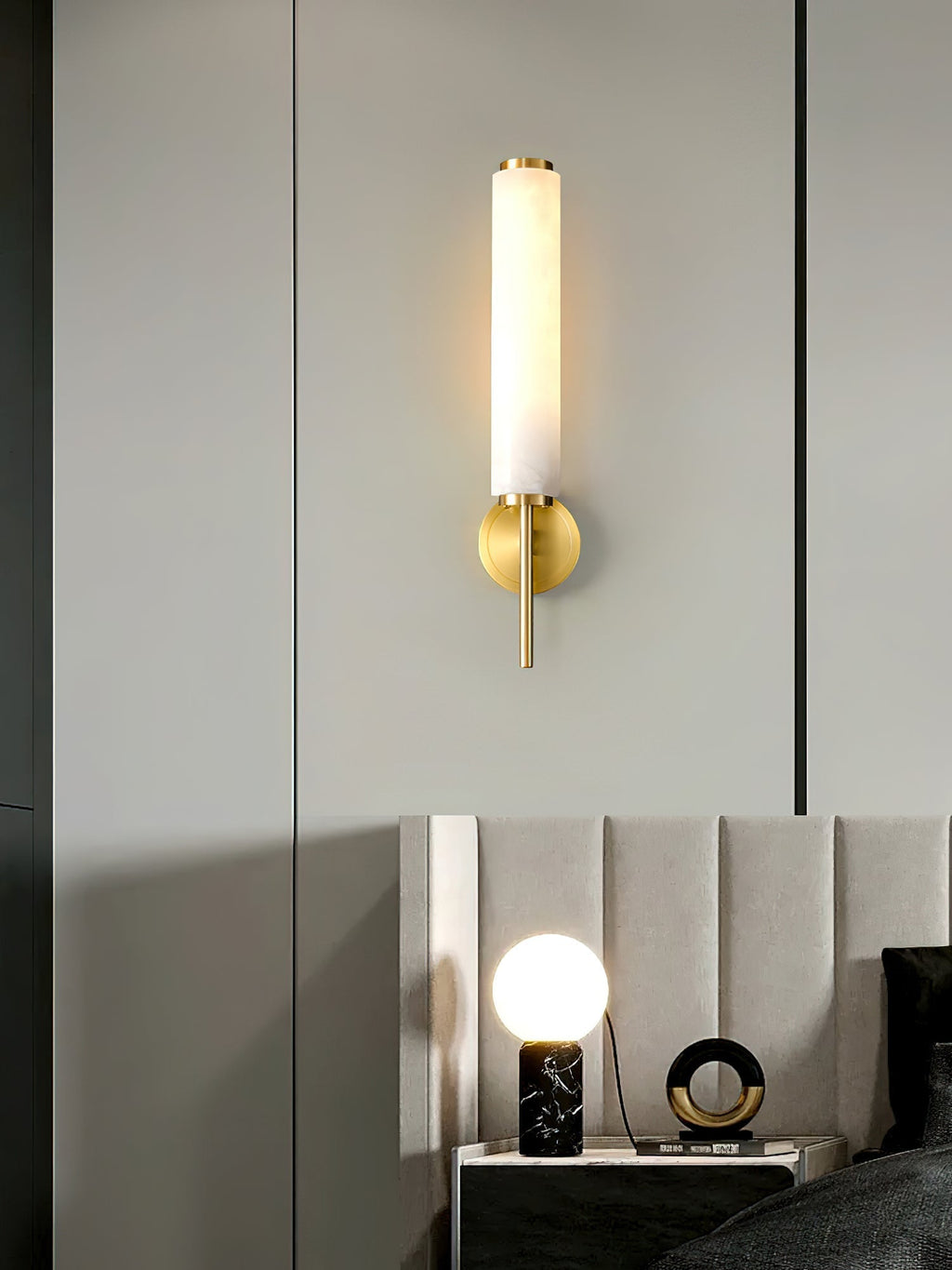 Clérachic Olrinyra in brass lit on a tall grey panel, with a bedroom reflected in the surface beside it.