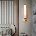 The Clérachic Olrinyra in brass lit on a fluted grey wall, beside a made bed and a small nightstand.