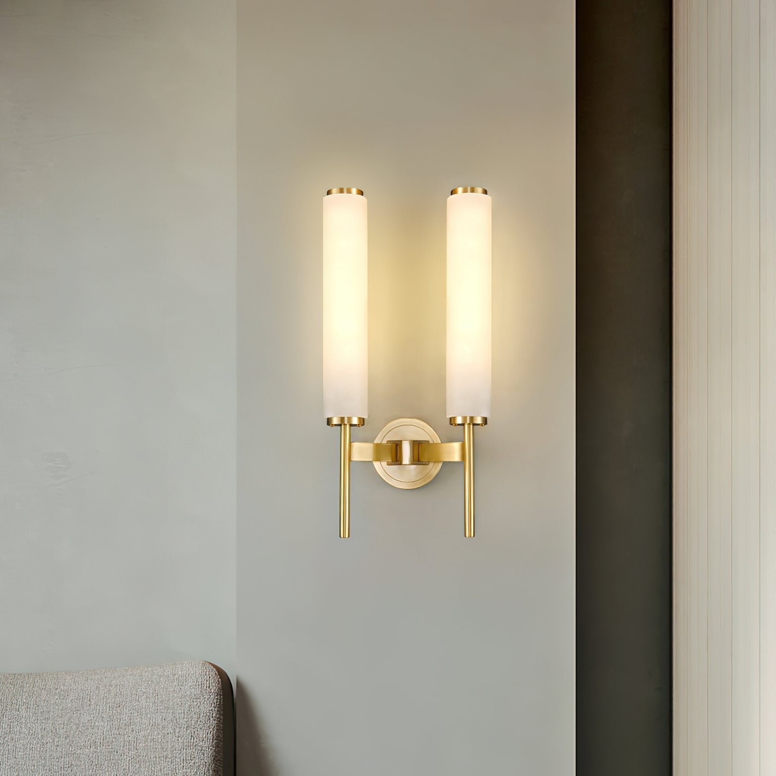 The Clérachic Olrinyra twin in brass lit on a pale wall, with a sofa arm just entering the lower frame.