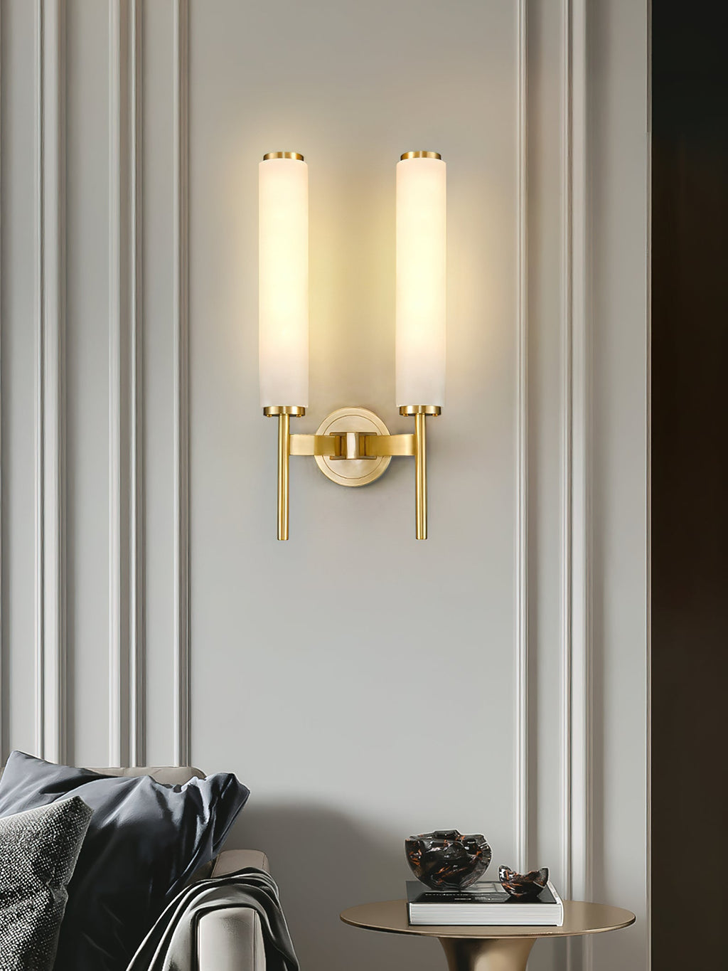 Clérachic Olrinyra twin in brass lit on a panelled wall, above a sofa cushion and a small round side table.