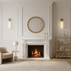 A pair of Clérachic wall lights lit either side of a round gold mirror above a white marble fireplace with a lit fire.