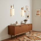 Two gold Clérachic wall lights with stacked opal balls on a cream wall, above a walnut sideboard with framed prints and a patterned rug.