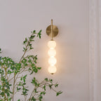 Gold Clérachic wall light lit against a pale pink wall, with a leafy branch crossing in front of its four opal balls.