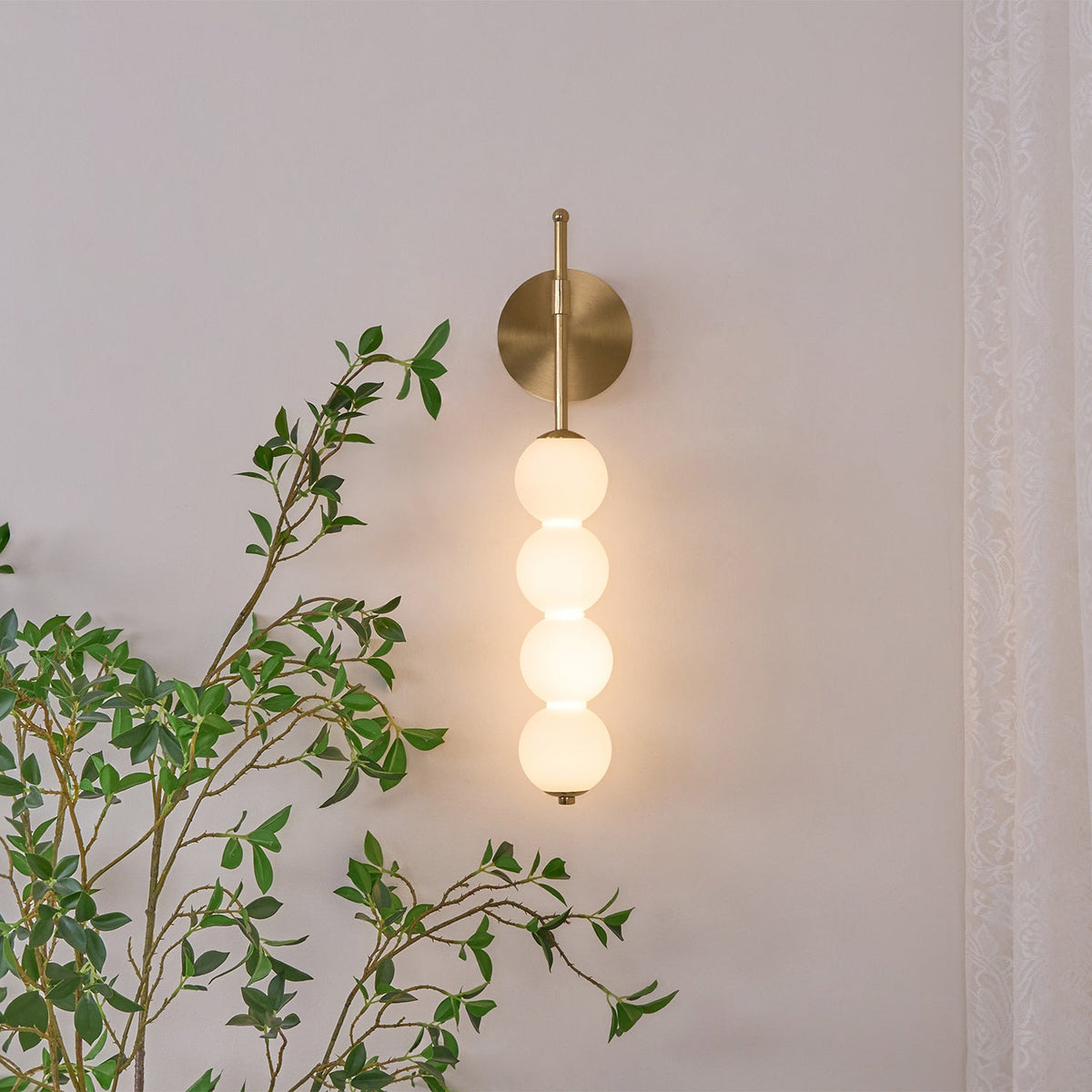Gold Clérachic wall light lit against a pale pink wall, with a leafy branch crossing in front of its four opal balls.