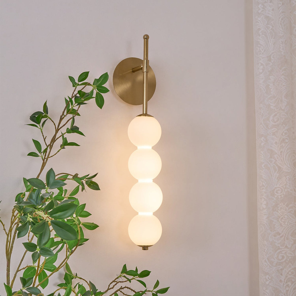 Close view of the lit gold Clérachic wall light, its four opal balls glowing warm beside a leafy green branch and lace curtain.