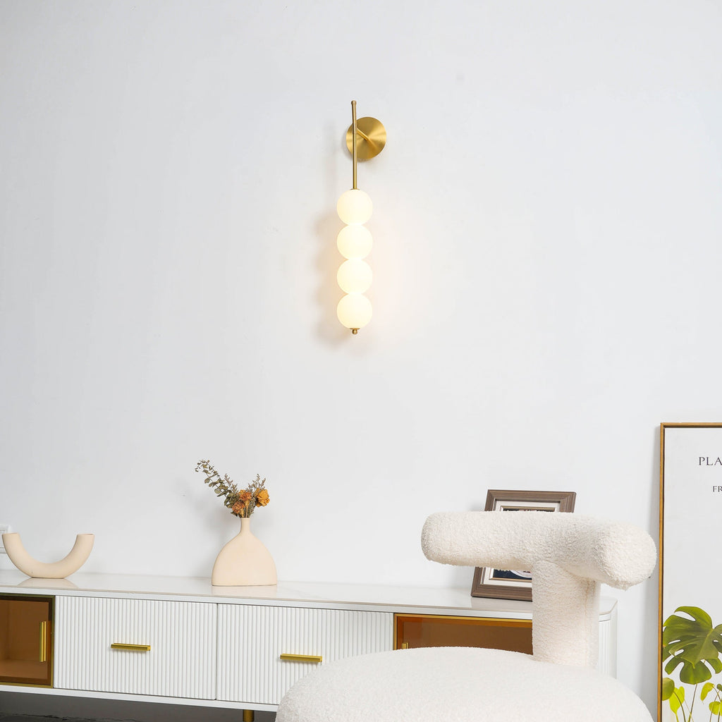 Gold Clérachic wall light on a white wall above a ribbed white sideboard, with a cream vase, a boucle chair and a framed print.