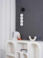 Black-finish Clérachic wall light with unlit white balls on a grey wall, above a white arched shelving unit with a small vase and books.