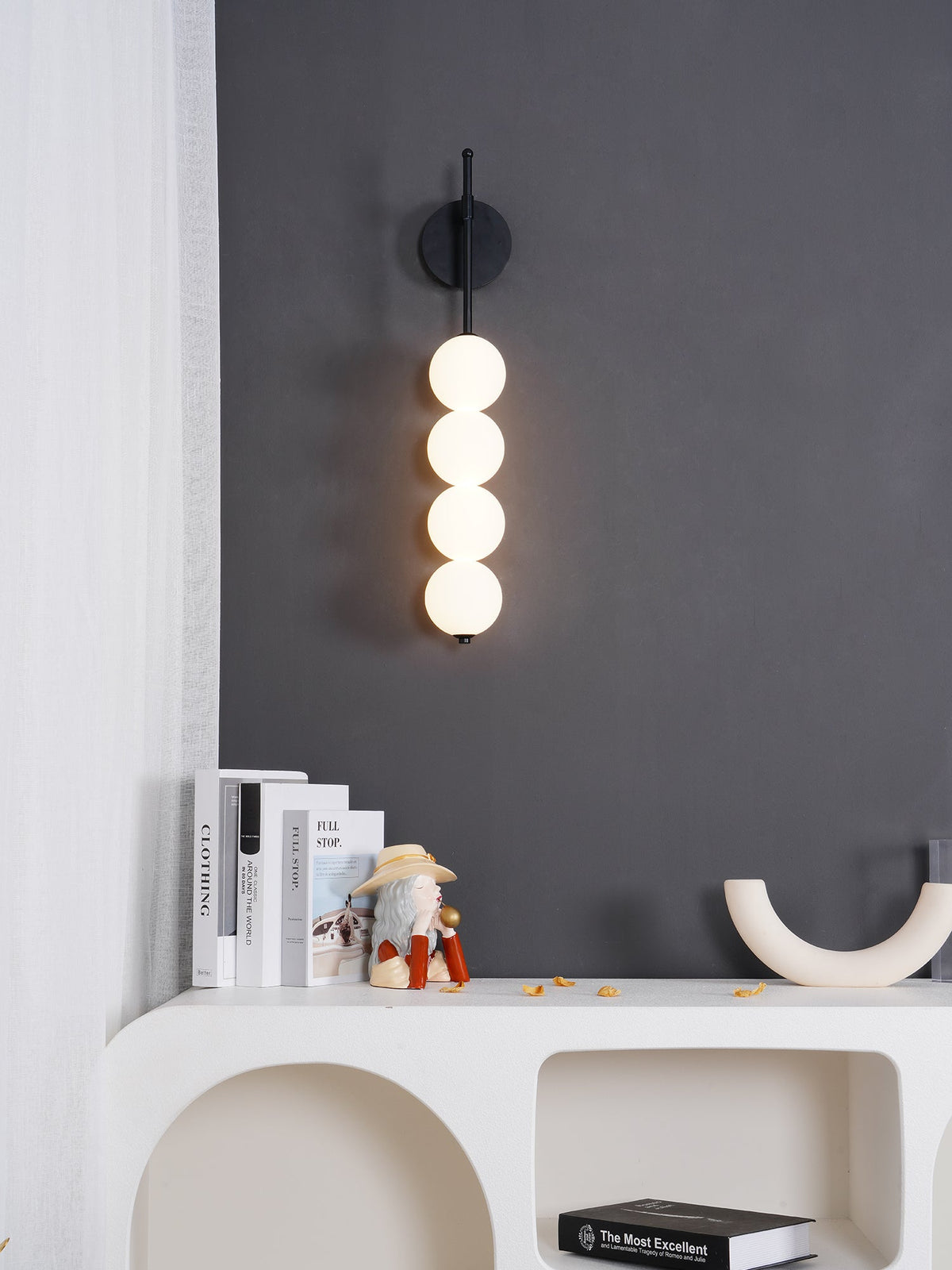 Black-finish Clérachic wall light glowing on a charcoal wall above a white arched console, books and a cream U-shaped ornament.