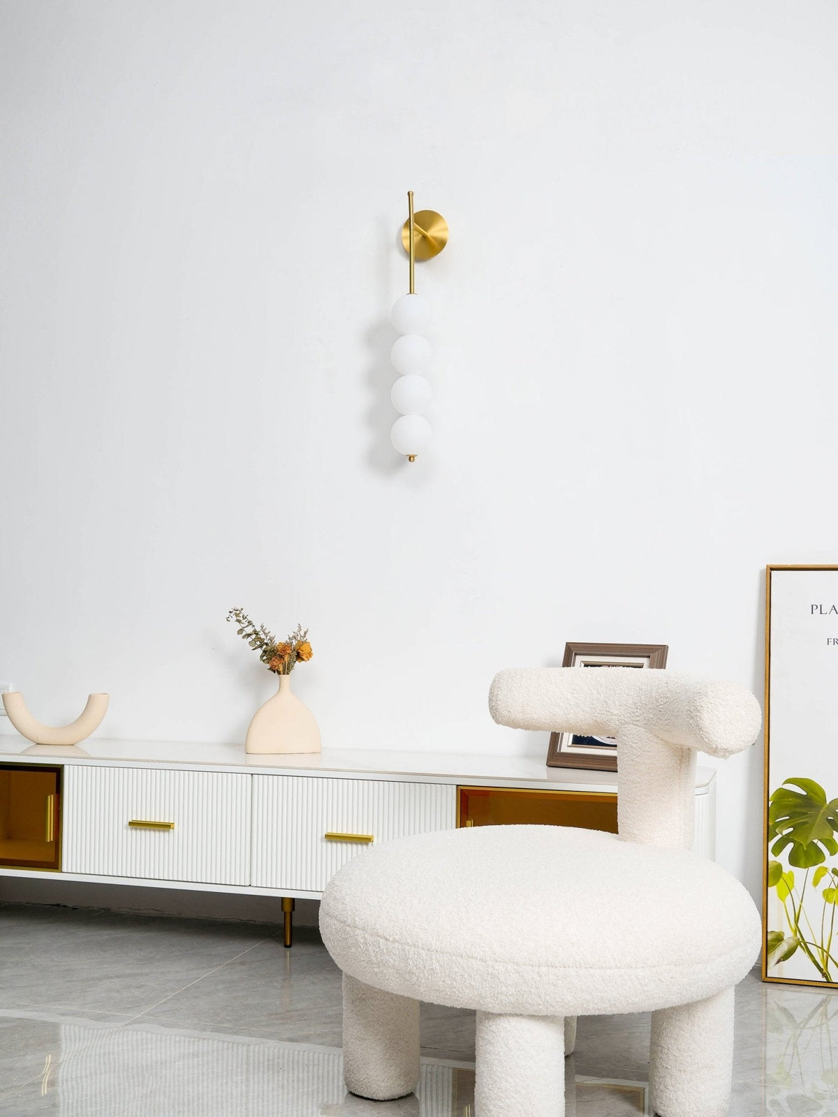 Gold Clérachic wall light, unlit, high on a white wall above a long ribbed sideboard and a white boucle stool.