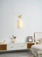 Gold Clérachic wall light, lit, on a white wall above a low ribbed sideboard with a cream vase and a framed picture.