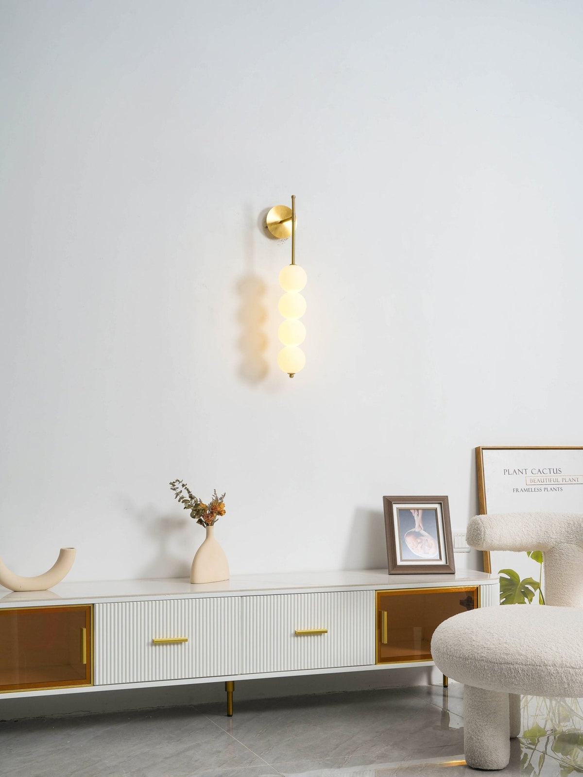 Gold Clérachic wall light, lit, on a white wall above a low ribbed sideboard with a cream vase and a framed picture.
