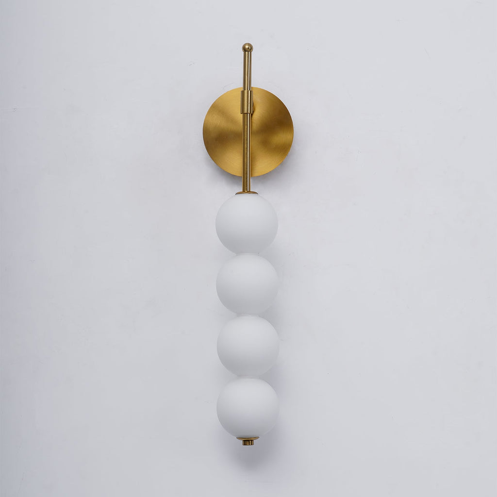 Close view of the gold Clérachic wall light unlit, its four matt white balls hanging from a brass arm on a pale grey wall.