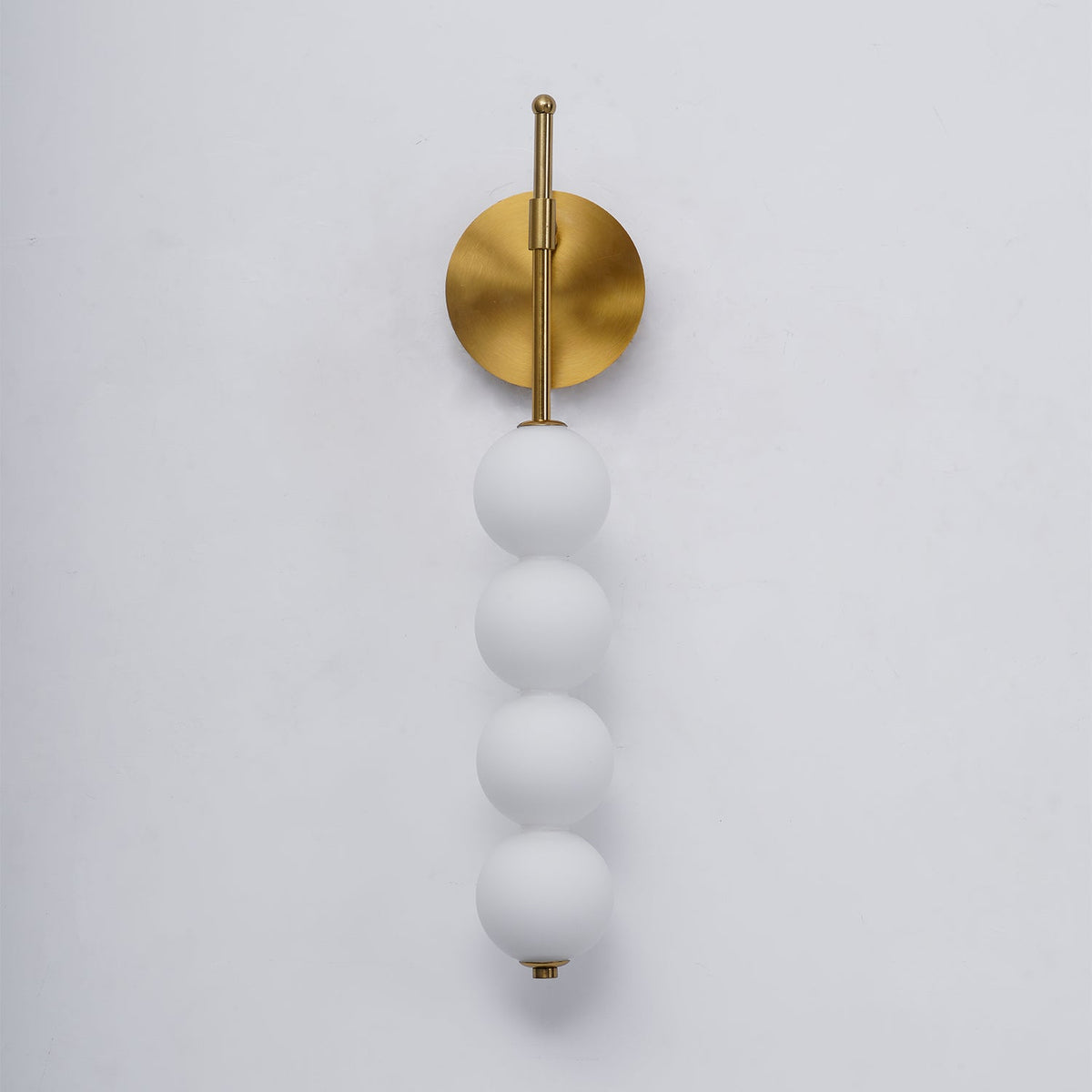 Close view of the gold Clérachic wall light unlit, its four matt white balls hanging from a brass arm on a pale grey wall.