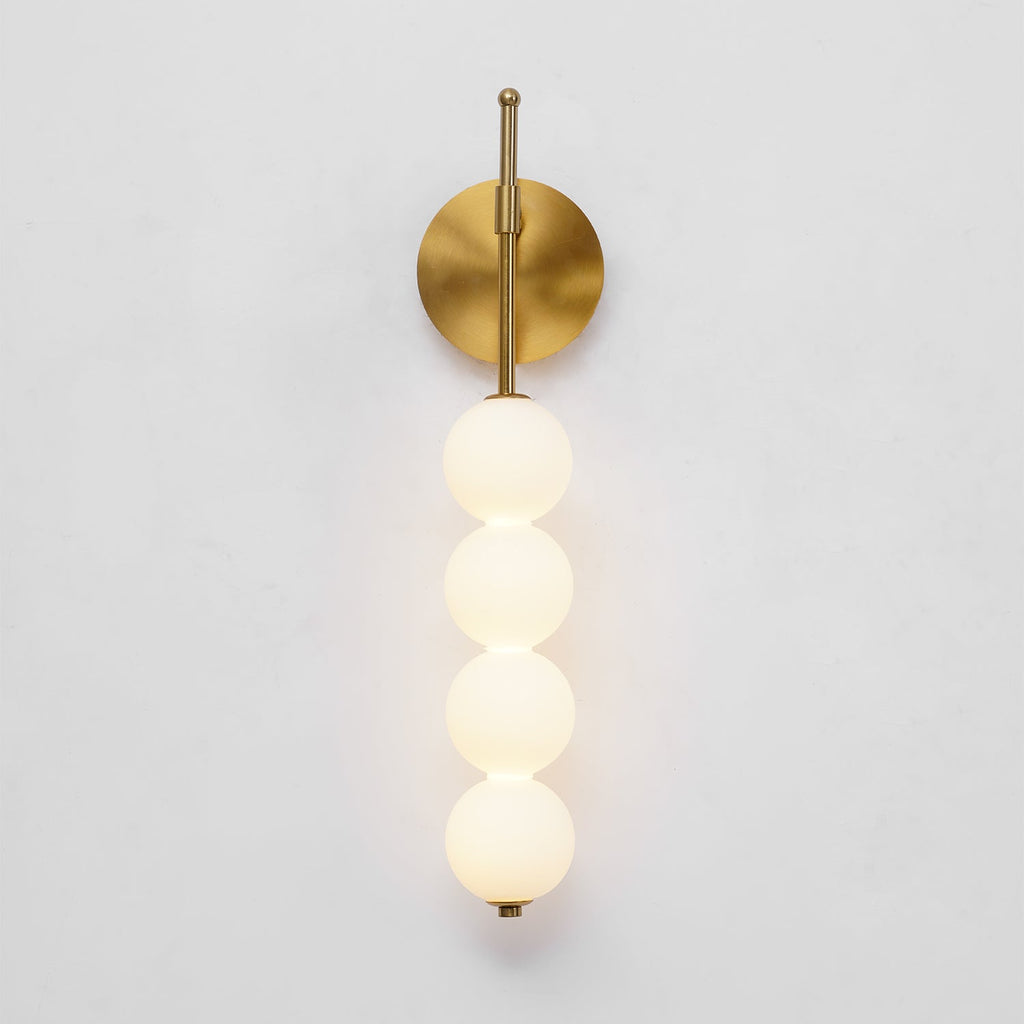 The gold Clérachic wall light lit on a pale grey wall, its four balls glowing a soft warm white.