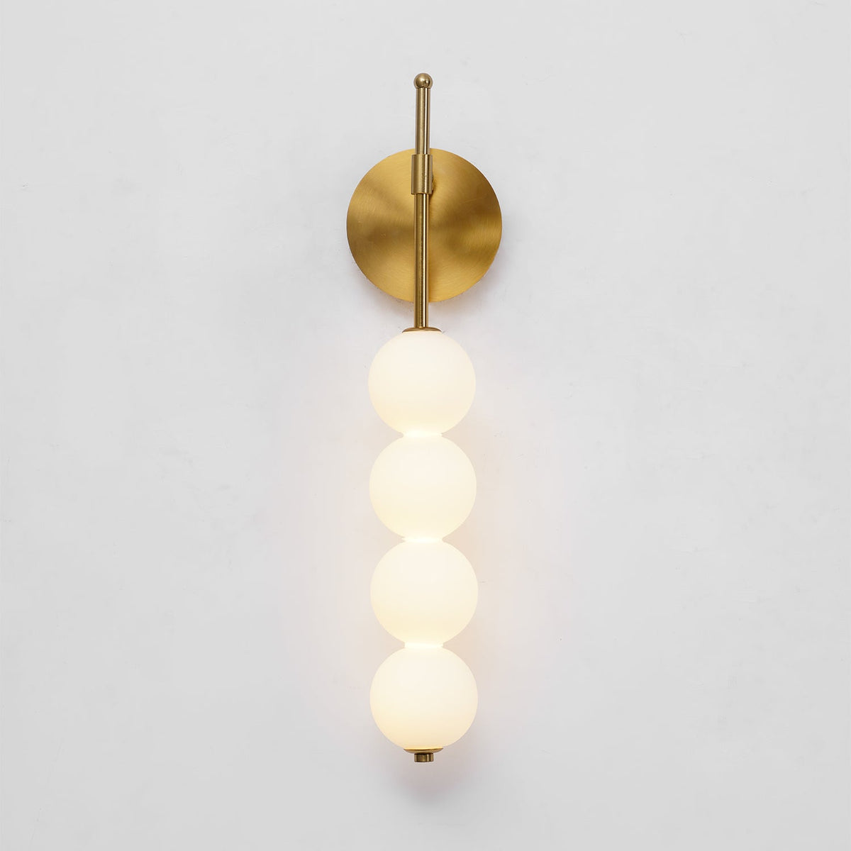 The gold Clérachic wall light lit on a pale grey wall, its four balls glowing a soft warm white.