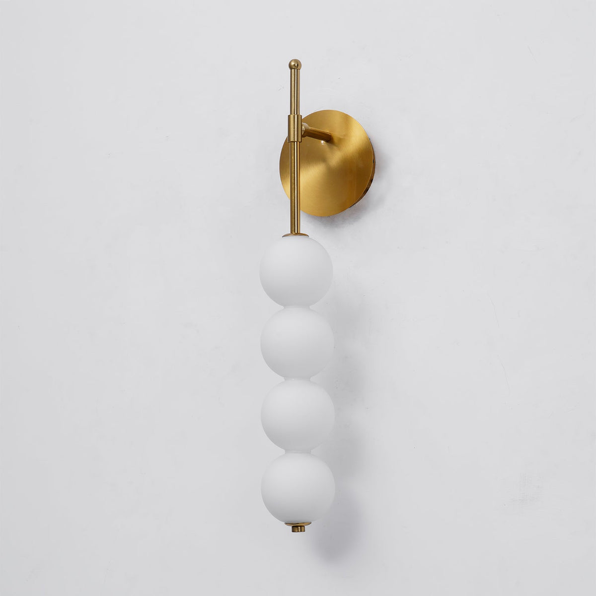 Gold Clérachic wall light, unlit, seen at a slight angle on a pale wall, with a soft shadow of the four white balls beside it.