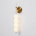 Angled view of the lit gold Clérachic wall light, the opal balls glowing and a warm reflection falling on the wall.
