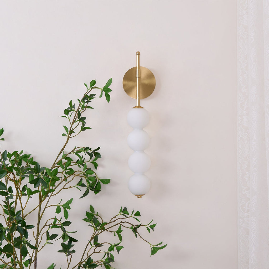 Gold Clérachic wall light, unlit, on a cream wall with a leafy branch crossing the foreground and a lace curtain at the right.