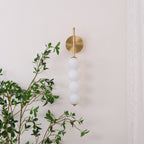 Gold Clérachic wall light, unlit, on a cream wall with a leafy branch crossing the foreground and a lace curtain at the right.