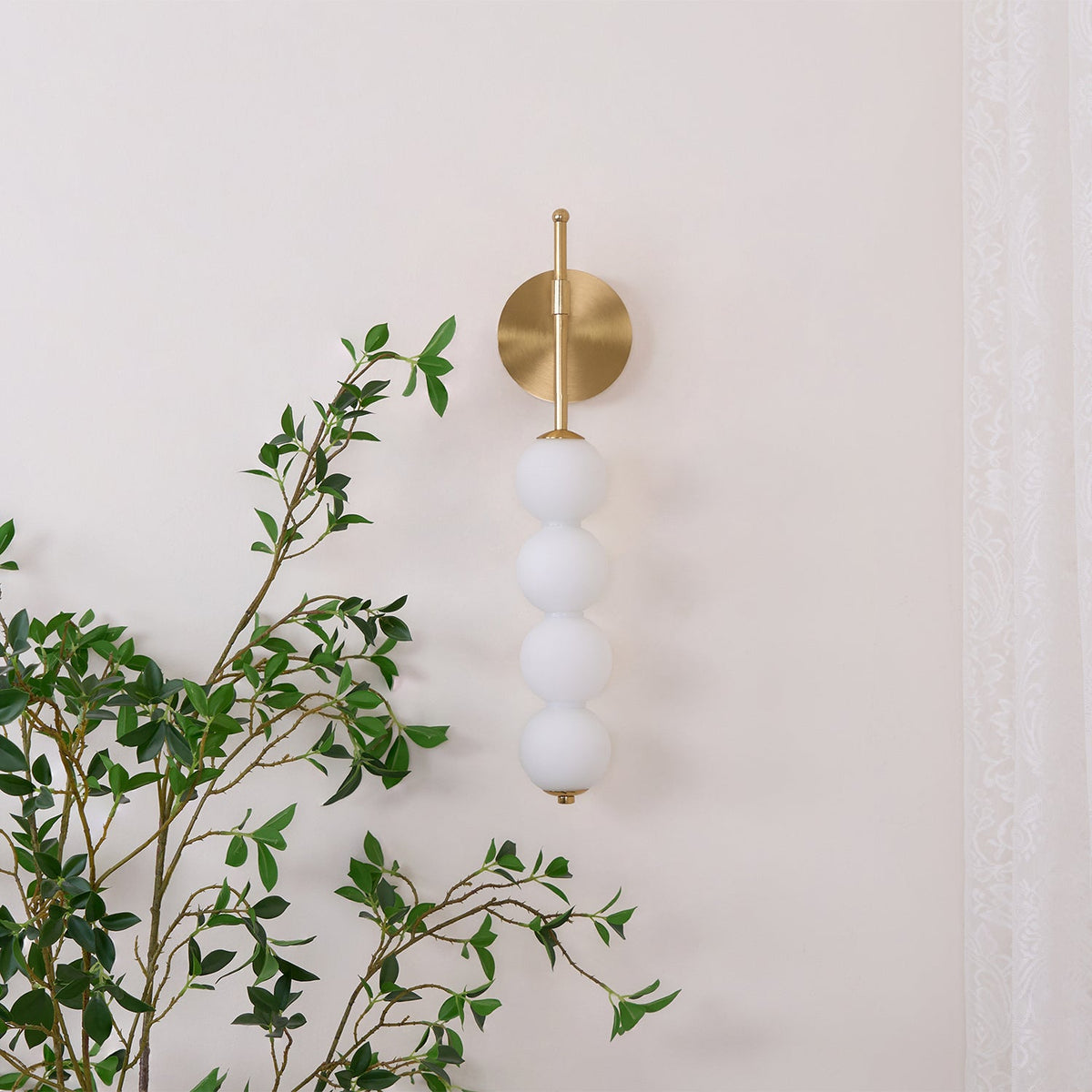 Gold Clérachic wall light, unlit, on a cream wall with a leafy branch crossing the foreground and a lace curtain at the right.