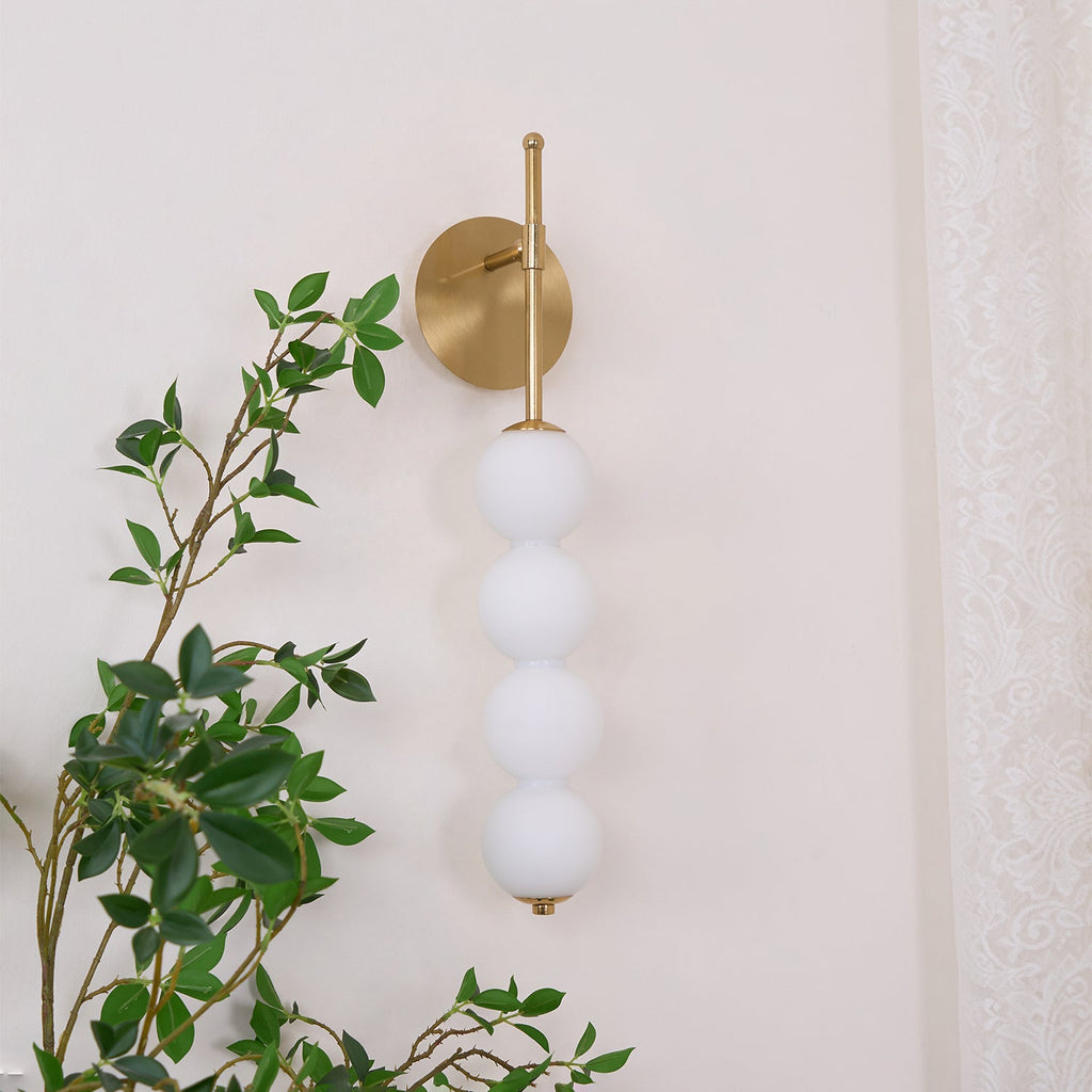 Gold Clérachic wall light with four matt white balls, unlit, beside a green leafy branch and a lace curtain.
