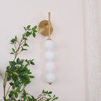 Gold Clérachic wall light with four matt white balls, unlit, beside a green leafy branch and a lace curtain.