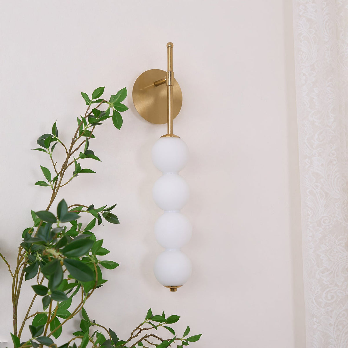 Gold Clérachic wall light with four matt white balls, unlit, beside a green leafy branch and a lace curtain.