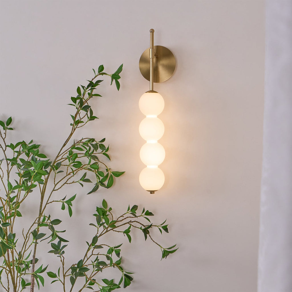 The Clérachic wall light lit in gold, the opal balls glowing warm among green leaves on a pale wall.