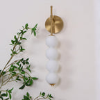 Gold Clérachic wall light, unlit, with four smooth white balls on a pale wall, and leafy branches at the left.