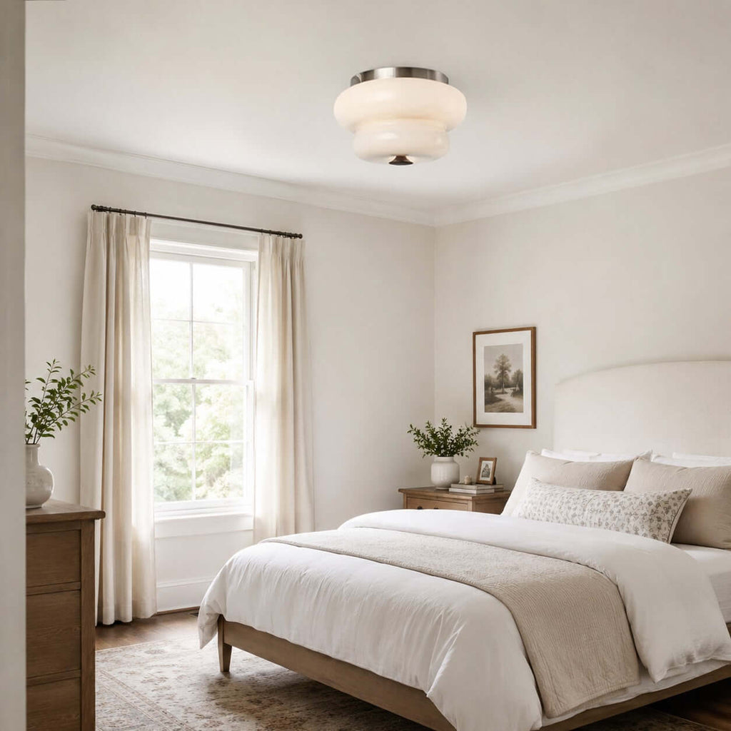 A Clérachic flush-mount light on the ceiling of a bedroom with a white bed, a tall window and linen curtains.