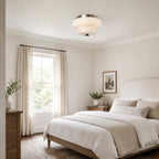 A Clérachic flush-mount light on the ceiling of a bedroom with a white bed, a tall window and linen curtains.