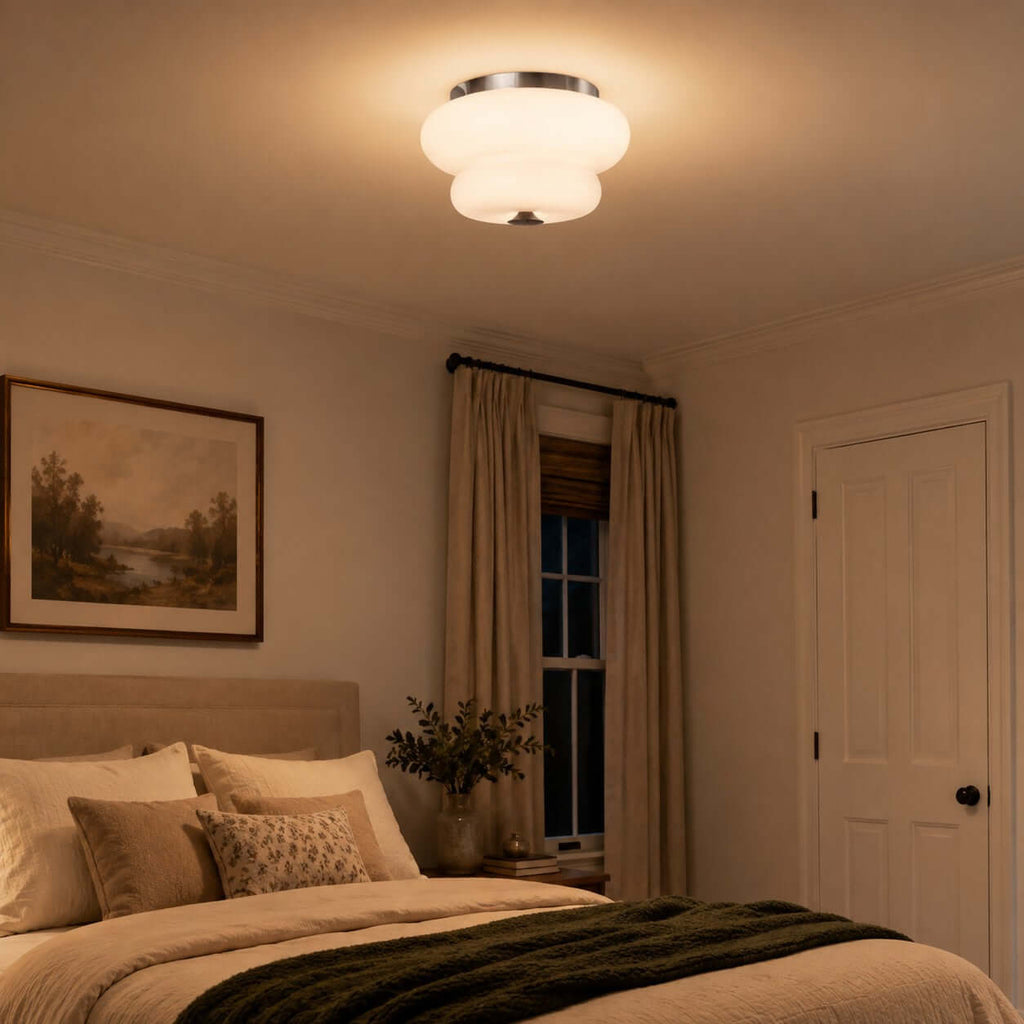 A Clérachic flush-mount light casts a warm glow over a bedroom with a green throw and a framed landscape painting.