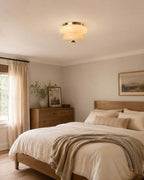 A Clérachic flush-mount light above a bed with a woven throw, a wooden dresser and a framed landscape print.