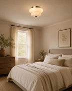 A Clérachic flush-mount light above a bedroom with a textured throw, a wooden dresser and a leafy plant by the window.