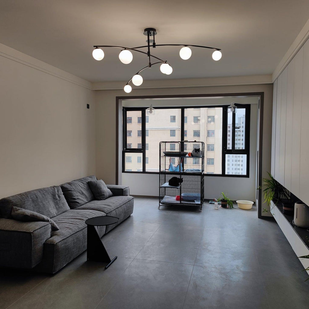Black Clérachic chandelier switched on in a wide grey living room, above a charcoal sofa and a tall window.