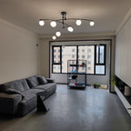 Black Clérachic chandelier switched on in a wide grey living room, above a charcoal sofa and a tall window.