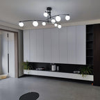 Black Clérachic chandelier with frosted globes in a living room with white wall cabinets and a low shelf of plants.
