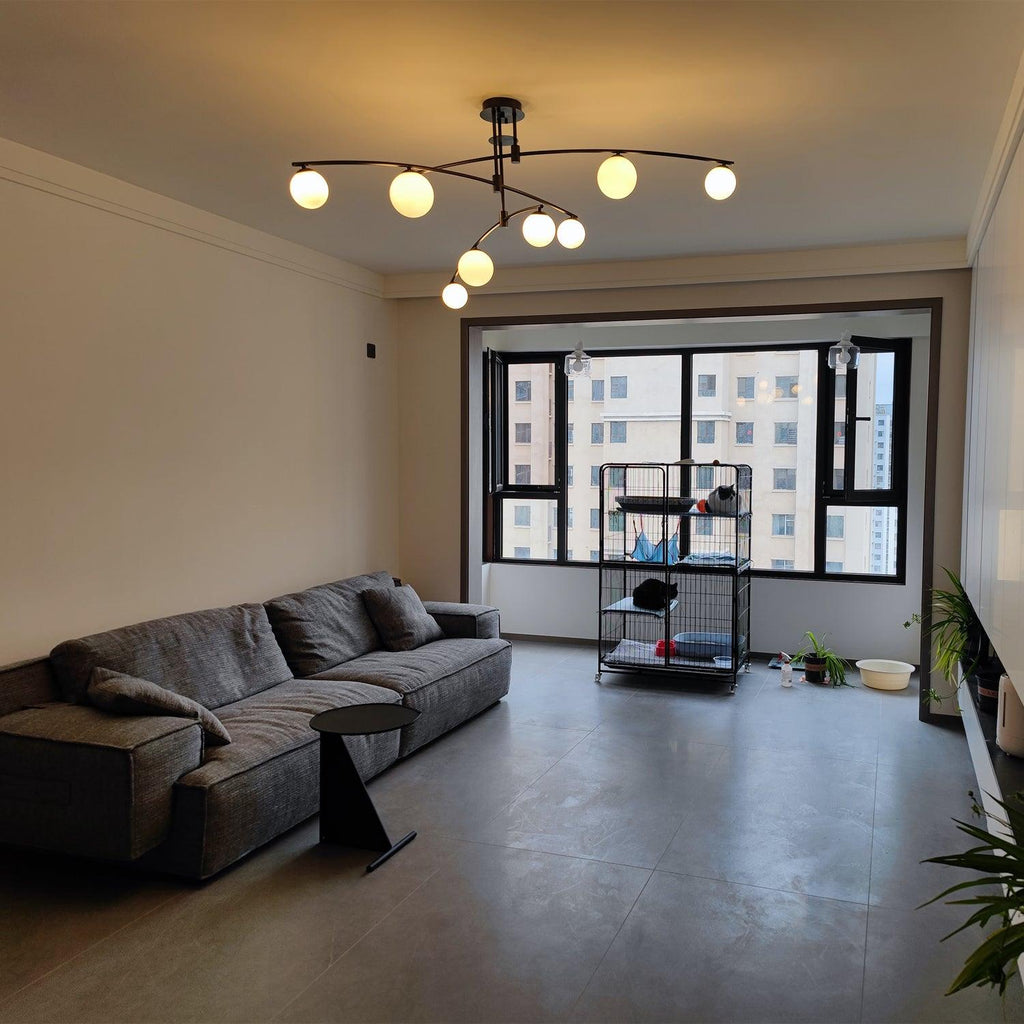 This Clérachic black chandelier glowing a warmer, yellower white in a grey living room with a sofa and a tall window.